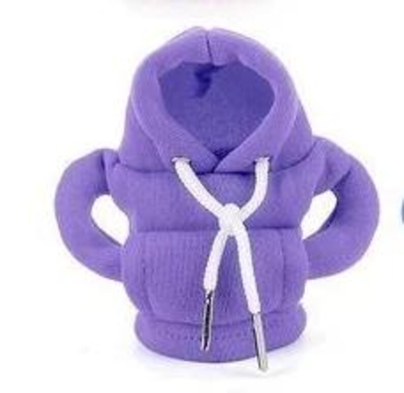 May include: A small, lavender-colored hoodie with a hood and sleeves. The hoodie has a white drawstring with silver-colored aglets. The fabric appears to be a soft, textured material.