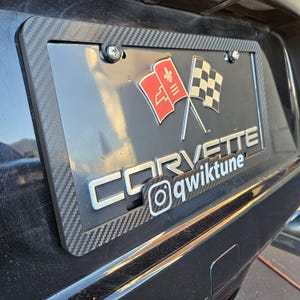 May include: A carbon fiber license plate frame with a black background. The frame features the word "CORVETTE" in silver lettering, along with the Instagram handle "@qwiktune". The license plate has red and checkered flags.