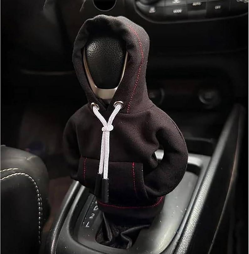 May include: A black hoodie-shaped car gear shift cover with red stitching and a white drawstring. The cover is designed to fit over the gear shift knob, adding a unique and stylish touch to the car's interior.