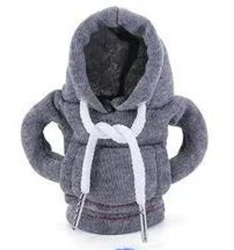 May include: A gray hoodie-shaped bottle cooler with white drawstrings. The cooler has a hood and sleeves, designed to fit around a bottle. The drawstrings are tied at the neck. The cooler is made of a soft, textured fabric.