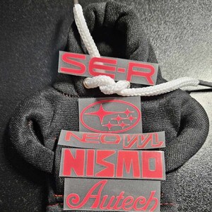 May include: Miniature black hoodie with white drawstring and red text decals. The decals include "SE-R", a Subaru logo, "NEOVL", "NISMO", and "Autech". The hoodie is made of a textured fabric.