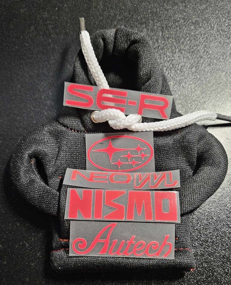 May include: Miniature black hoodie with white drawstring and red text decals. The decals include "SE-R", a Subaru logo, "NEOVL", "NISMO", and "Autech". The hoodie is made of a textured fabric.