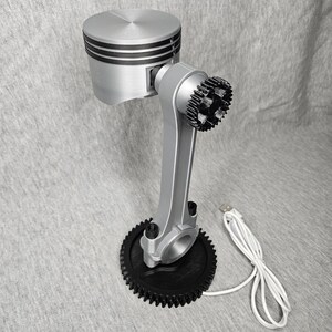 May include: A silver and black desk lamp crafted from engine components. The lamp features a piston head, connecting rod, and gear-shaped base. A USB cable is attached for power. The lamp is a unique piece of industrial decor.