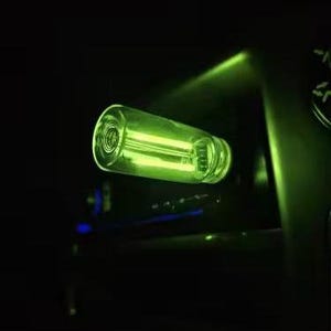 May include: A cylindrical glass object with a green internal light source. The object has a ridged design on the end and is set against a dark background. The light emits a bright, neon green glow.