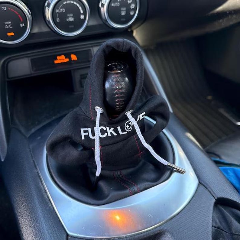 May include: Black shift knob cover with a hoodie design. The hoodie has white drawstrings and the words "FUCK LOVE" printed in white. The cover is designed to fit over a car's gear shift.
