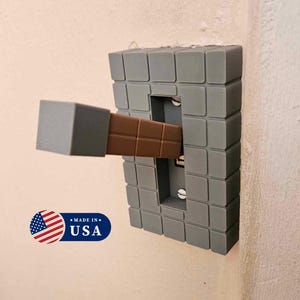 May include: A gray light switch cover with a brown lever sticking out of it. The cover is made to look like a stone block from the video game Minecraft. The lever is made to look like a Minecraft pickaxe. The text "MADE IN USA" is printed on a small American flag sticker below the switch.