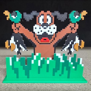 May include: A 3D-printed sculpture of the Duck Hunt dog holding two pixelated ducks. The dog is brown with black ears and a white muzzle. The ducks are black, white, green, and orange. Green pixelated grass is at the base.