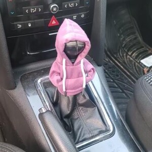 May include: A small, purple hoodie with white drawstrings, placed on a car's gear shift. The hoodie is a solid color and appears to be made of a soft material. The car's interior is visible in the background, including the dashboard and center console.