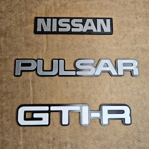 Puede incluir: Tres insignias de emblema de coche: "NISSAN" en plata sobre fondo negro, "PULSAR" en plata con contorno negro y "GT-R" en blanco con contorno negro. Las insignias están dispuestas verticalmente sobre una superficie marrón claro.