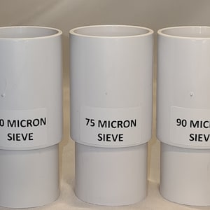 Może przedstawiać: Trzy białe plastikowe sita z etykietami wskazującymi ich rozmiar w mikronach: sito 50 mikronów, sito 75 mikronów i sito 90 mikronów.
