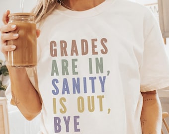 May include: Cream-colored t-shirt with the text "GRADES ARE IN, SANITY IS OUT, BYE" in colorful letters. The shirt is worn with white socks with black stripes. A glass of iced coffee is held in the hand.