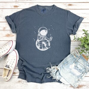 May include: A dark blue t-shirt with a white graphic of an astronaut sitting on the moon and eating ice cream. The astronaut is wearing a helmet and a spacesuit. There are stars in the background.