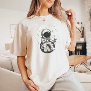 May include: A white t-shirt with a black and white illustration of an astronaut sitting on a planet and holding an ice cream cone. The astronaut is wearing a space suit and has a helmet on. There are small stars around the astronaut.