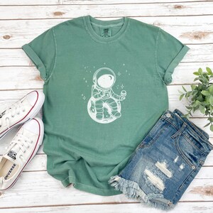 May include: A green t-shirt with a white graphic of an astronaut sitting on the moon and eating ice cream. The astronaut is wearing a helmet and a spacesuit. There are stars in the background.