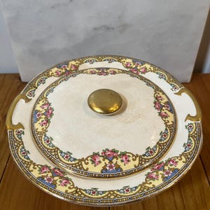 May include: A vintage white porcelain covered dish with a floral design in shades of pink, blue, and yellow. The dish has a gold trim and a gold knob on the lid.