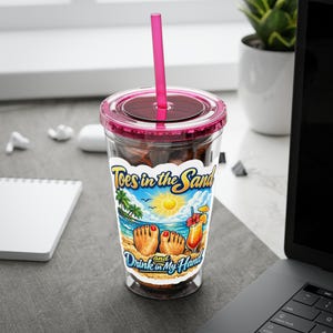 May include: A clear plastic tumbler with a pink lid and straw. The tumbler features a colourful beach scene with the text "Toes in the Sand and Drink in My Hand". The cup is filled with ice and a dark liquid.