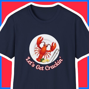 May include: A navy blue t-shirt featuring a white plate with a red lobster on it. The text "Let's Get Crackin'" is written in white below the plate.