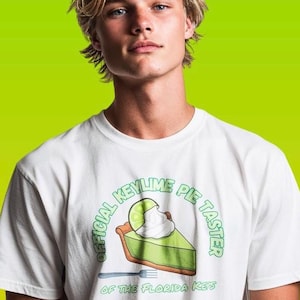 Puede incluir: Camiseta blanca con un gráfico de una porción de tarta de lima y el texto "OFFICIAL KEY LIME PIE TASTER OF THE FLORIDA KEYS" en verde. La camiseta tiene cuello redondo y mangas cortas.