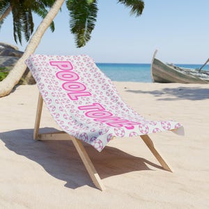 Puede incluir: Una toalla de playa blanca con un estampado rosa de salvavidas y las palabras "POOL TIME" en letras rosas grandes. La toalla está sobre una silla de playa de madera en una playa de arena.