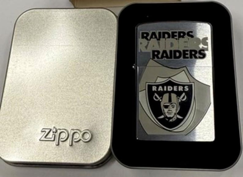 May include: A silver Zippo lighter with the Raiders logo and team name. The lighter is in a black case, and the lid of the silver Zippo case is open. The Zippo logo is visible on the case.