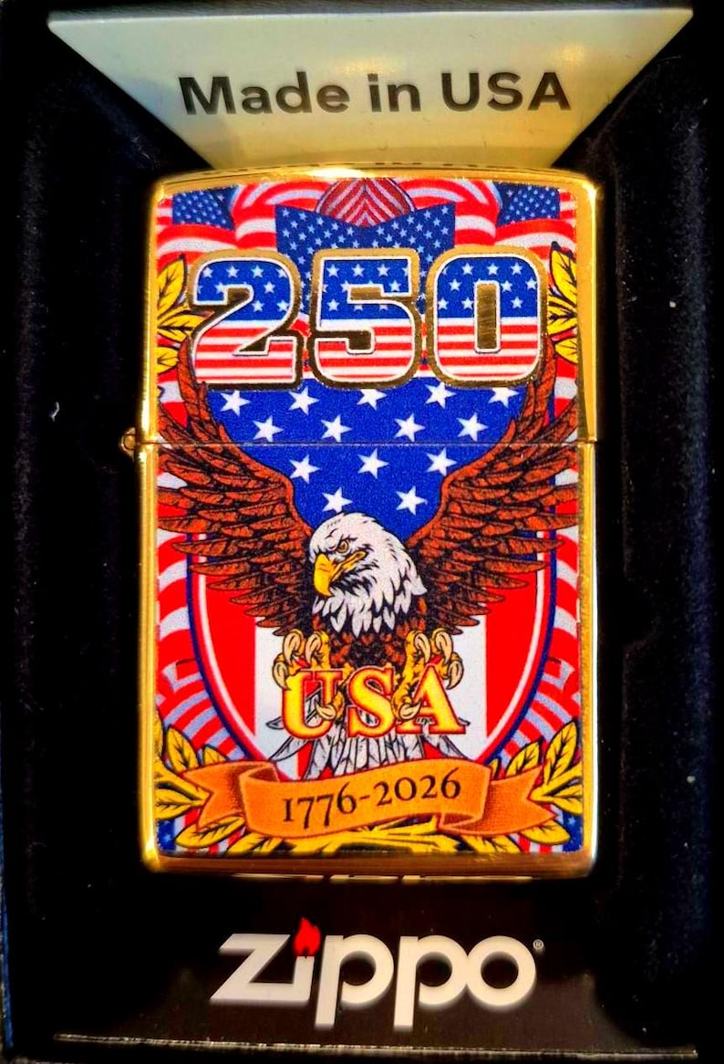 Puede incluir: Un encendedor Zippo dorado con un dise&ntilde;o patri&oacute;tico. El encendedor presenta el n&uacute;mero "250" en rojo y azul, un &aacute;guila con las alas extendidas y la palabra "USA". Tambi&eacute;n se muestra el texto "1776-2026".