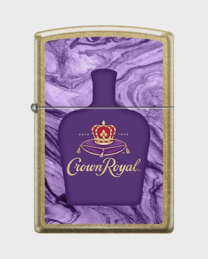 May include: A gold-toned lighter featuring a purple bottle design with a crown and the words "Crown Royal." The background has a marbled purple and white pattern. The lighter is rectangular and has a hinged lid.