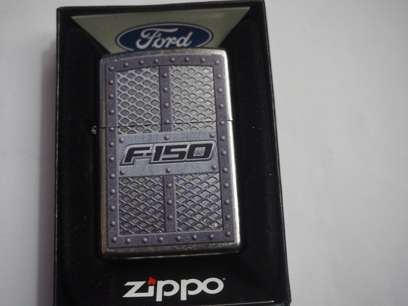 Puede incluir: Un encendedor Zippo plateado con un dise&ntilde;o de Ford F-150. El encendedor tiene un acabado de metal plateado con un patr&oacute;n de cuadr&iacute;cula negro y gris y las palabras "Ford" y "F-150" impresas en &eacute;l.