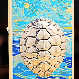 May include: A silver-coloured lighter with a raised turtle shell design. The background features a blue and yellow ocean and sky scene. The lighter has a rectangular shape with rounded edges.