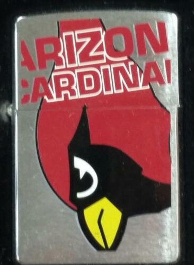 May include: A silver lighter with a red, white, and black Arizona Cardinals logo. The logo features a cardinal bird with a yellow beak and a white eye.