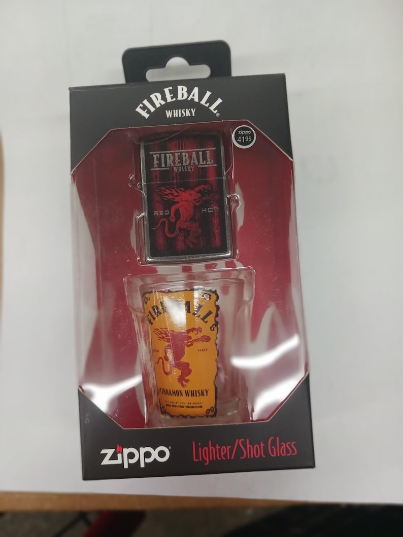 May include: A black box with a silver Zippo lighter and a shot glass featuring the Fireball Cinnamon Whisky logo. The lighter has a red and black design with the words "Fireball Whisky" and "Red Hot". The shot glass has a yellow and black design with the words "Fireball Cinnamon Whisky". The box has the words "Zippo Lighter/Shot Glass" printed on it.