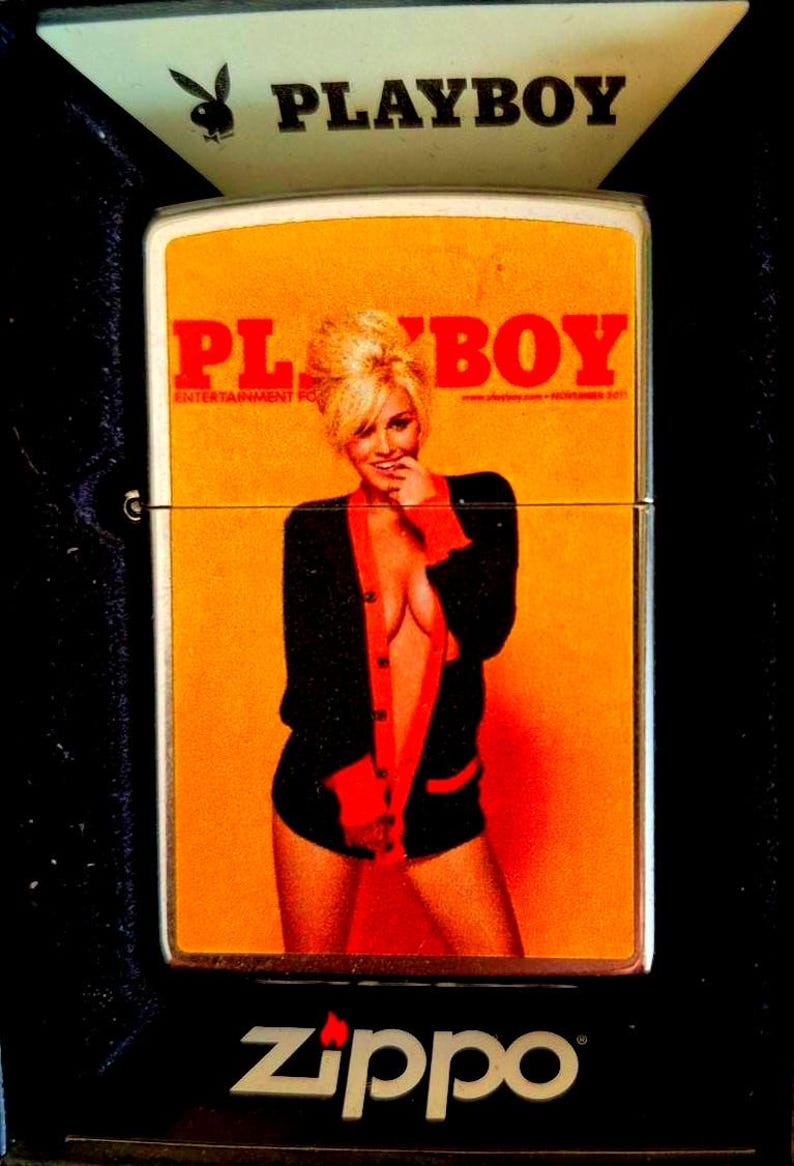 May include: A Zippo lighter with a Playboy magazine cover design. The lighter features a woman in a black and red cardigan against an orange background. The Playboy logo is at the top.