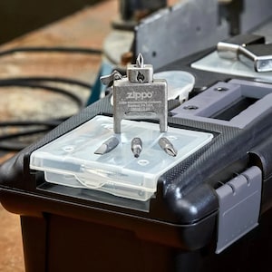 Puede incluir: Un kit multiherramienta Zippo plateado, con una herramienta en forma de encendedor y varias puntas de destornillador. El kit se muestra en una caja de herramientas negra con una bandeja de pl&aacute;stico transparente. El logotipo y la marca Zippo son visibles.