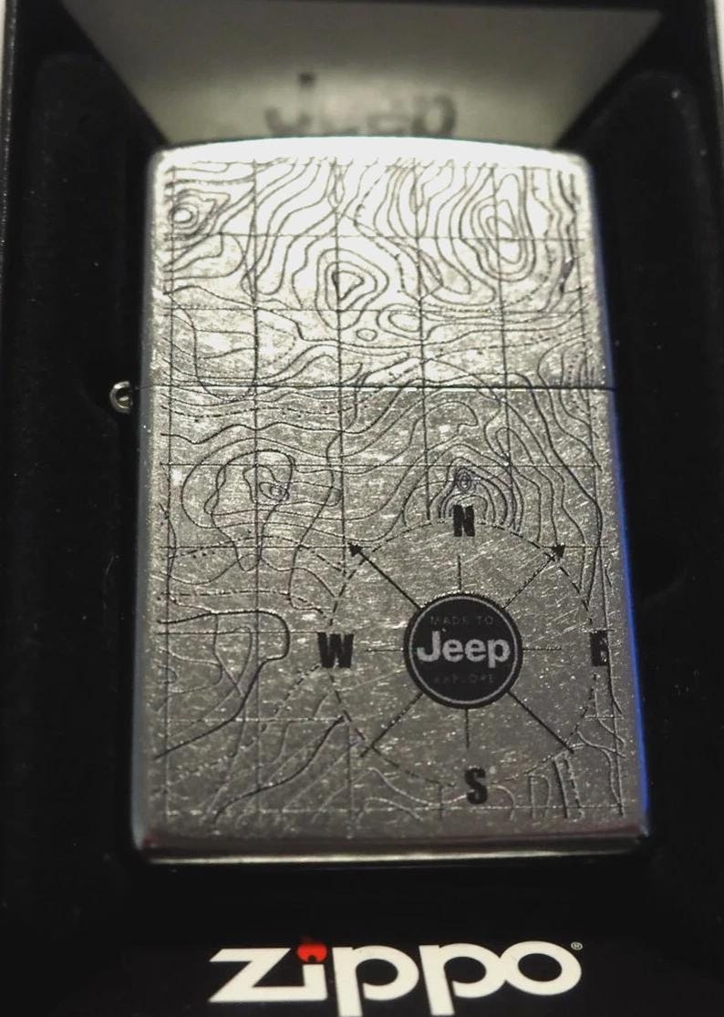 Op de afbeelding: Een zilveren Zippo-aansteker met een topografisch kaartontwerp. De aansteker heeft een kompasroos met de tekst "MADE TO Jeep EXPLORE". Het Zippo-logo is onderaan zichtbaar.