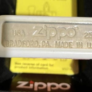 Op de afbeelding: Close-up van een Zippo-aansteker met de tekst "USA Zippo 250 BRADFORD, PA. MADE IN U.S.A." De aansteker is zilverkleurig met een witte rand. De achtergrond is geel en zwart.