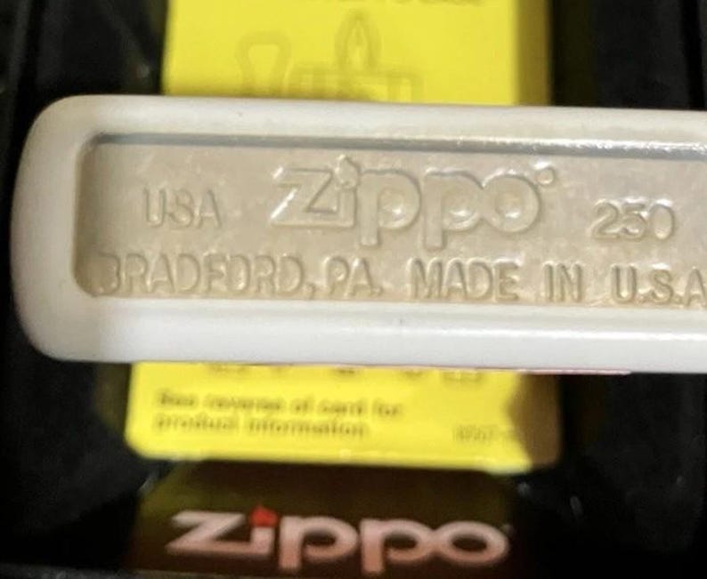Op de afbeelding: Close-up van een Zippo-aansteker met de tekst "USA Zippo 250 BRADFORD, PA. MADE IN U.S.A." De aansteker is zilverkleurig met een witte rand. De achtergrond is geel en zwart.
