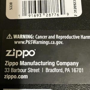 Op de afbeelding: Zwarte verpakking met de tekst "Genuine Zippo Lighter Made in USA". Een barcode label geeft "RED TOP/WHITE BOTTON" aan. Het Zippo-logo en bedrijfsinformatie zijn ook zichtbaar, samen met een waarschuwing over kanker en reproductieve schade.