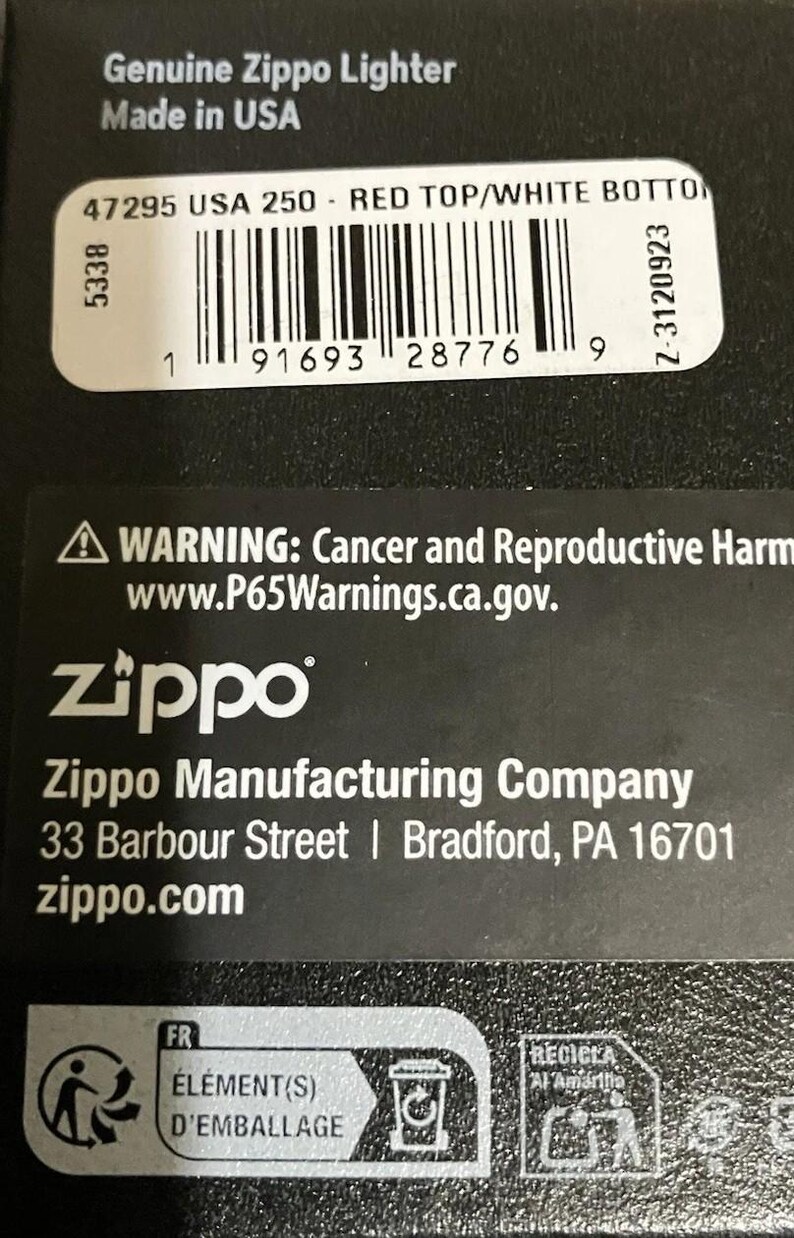Op de afbeelding: Zwarte verpakking met de tekst "Genuine Zippo Lighter Made in USA". Een barcode label geeft "RED TOP/WHITE BOTTON" aan. Het Zippo-logo en bedrijfsinformatie zijn ook zichtbaar, samen met een waarschuwing over kanker en reproductieve schade.