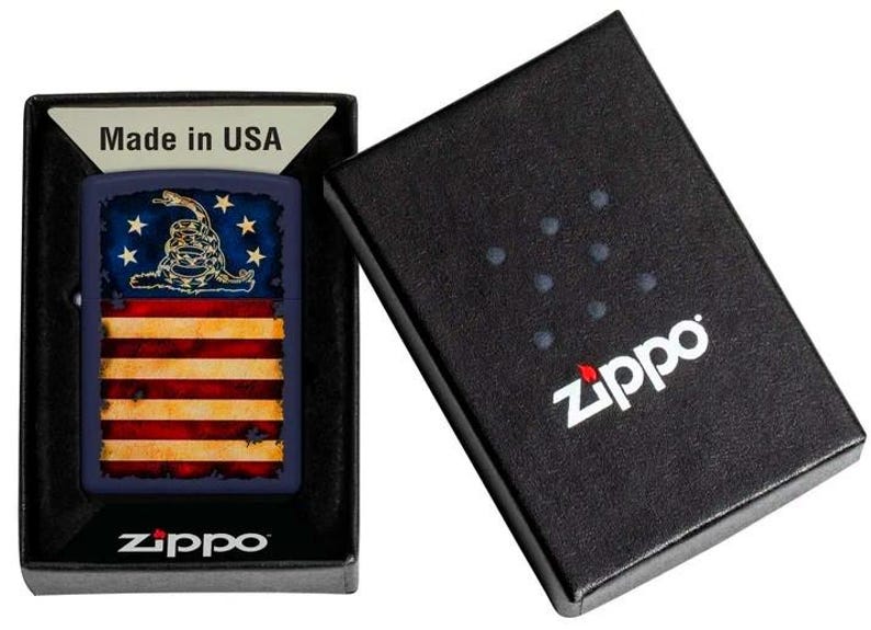 Puede incluir: Un encendedor de metal rectangular con un dise&ntilde;o de bandera de Gadsden con una serpiente de cascabel y el texto Don't Tread On Me, presentado en una caja negra de la marca Zippo con un inserto Made in USA. Acabado en azul, rojo y blanco.