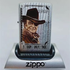 May include: A silver Zippo lighter with a portrait of John Wayne, the text "1969", and the name "JOHN WAYNE". The lighter is on a silver stand with the Zippo logo.