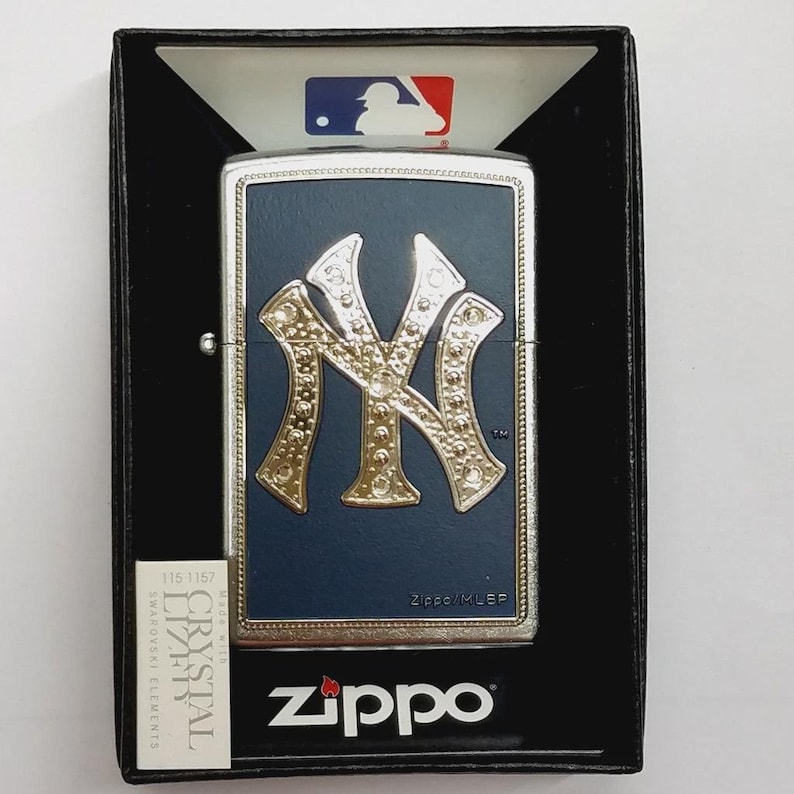 May include: A Zippo lighter in a black box. The lighter has a navy blue background with a gold-colored New York Yankees logo. The lighter is silver-colored around the edges. The box has the MLB logo.