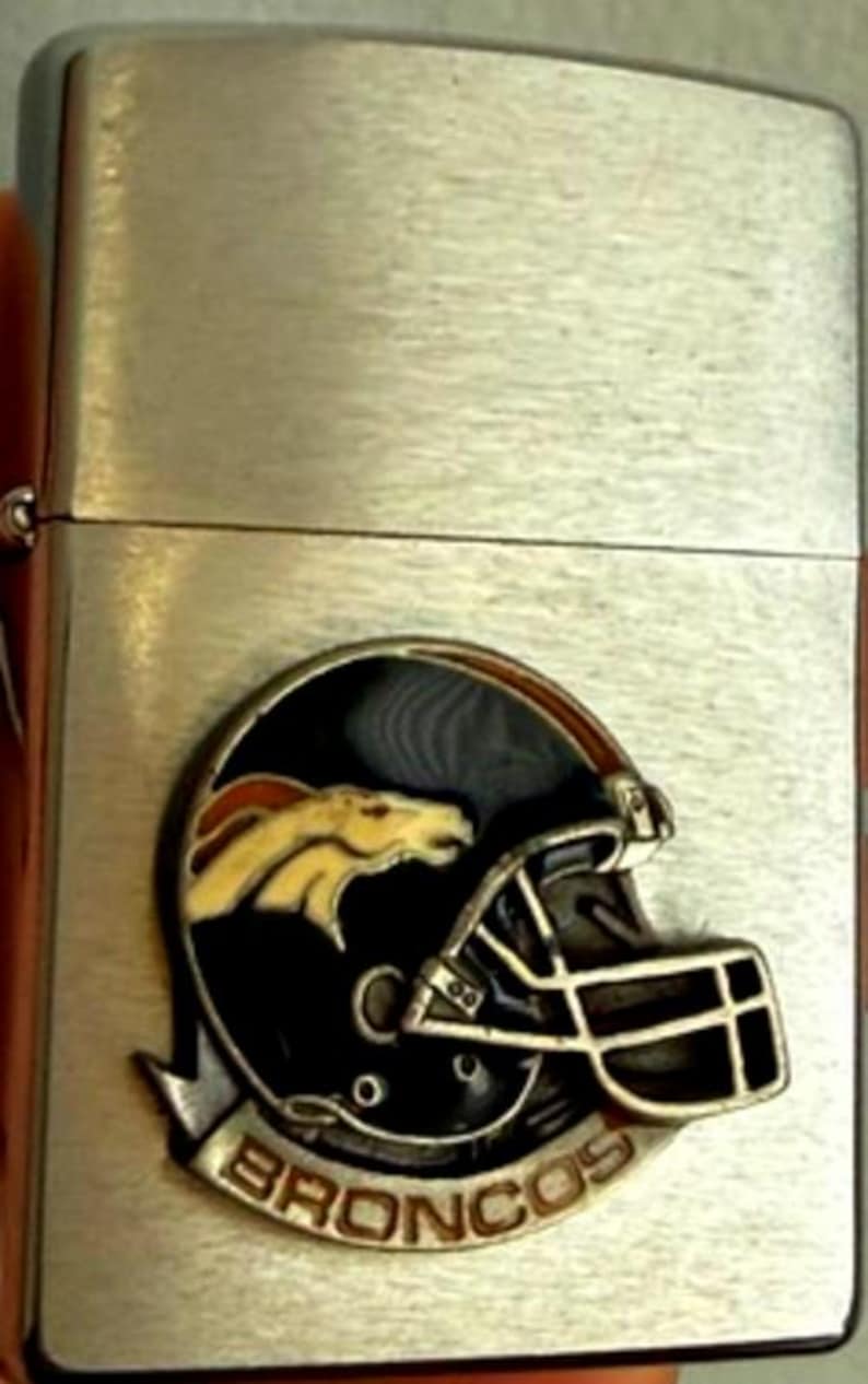 May include: A silver-colored lighter with a brushed metal finish. The lighter features a black, orange, and yellow emblem of a football helmet with the word "BRONCOS" at the bottom. The lighter is rectangular and has a hinged lid.