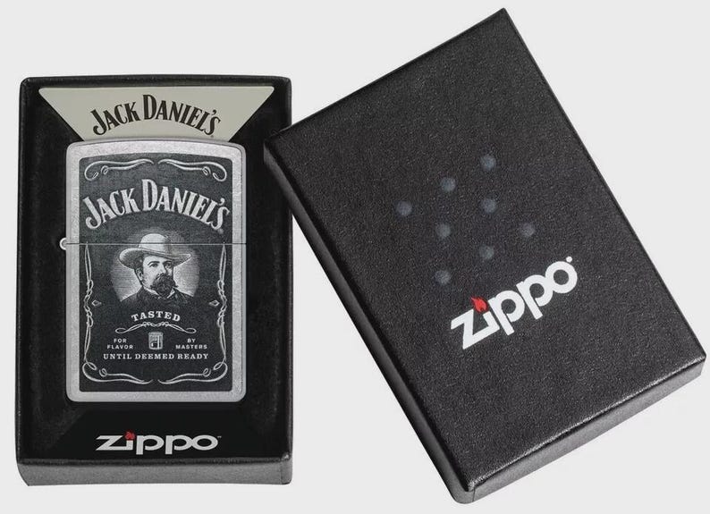 May include: A silver Zippo lighter with a Jack Daniel's label, featuring a portrait of a man in a hat. The lighter is in a black box with the Zippo logo. The box is open, revealing the lighter.