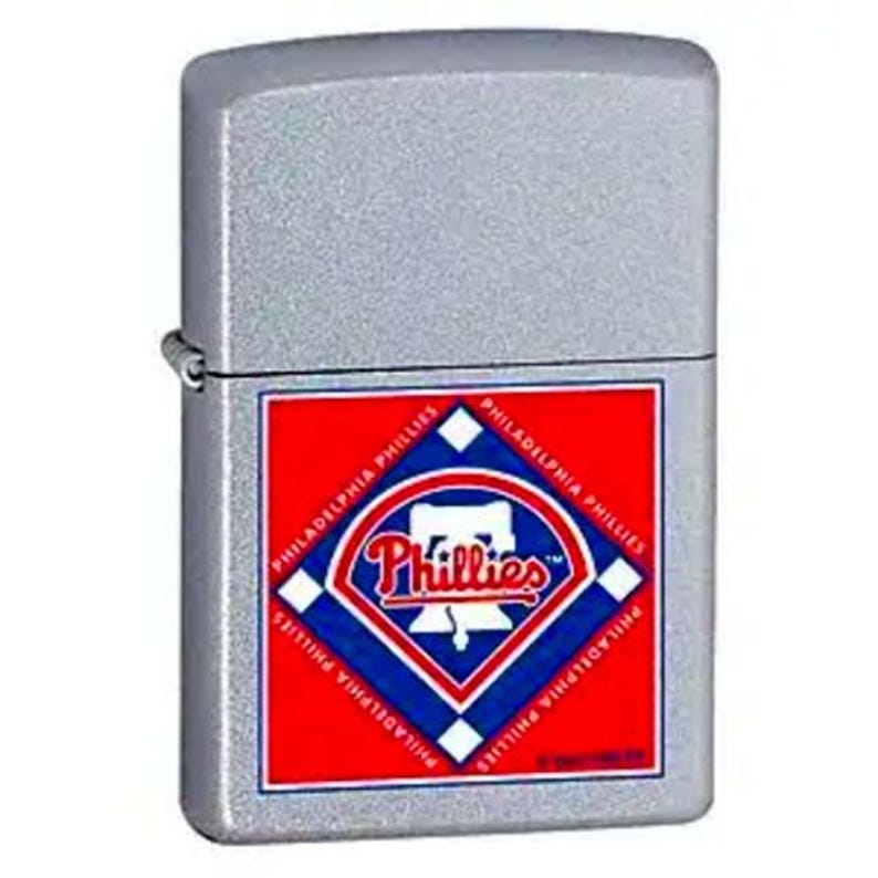 Puede incluir: Un encendedor plateado de forma rectangular. El encendedor presenta un emblema rojo, blanco y azul con la palabra "Phillies" y las palabras "Philadelphia Phillies" alrededor del emblema. El encendedor tiene una superficie texturizada.