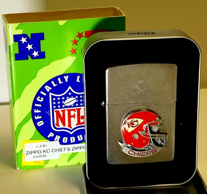 May include: A silver Zippo lighter with a red and white Kansas City Chiefs helmet emblem, presented in a black metal case. The box is green with an NFL logo and the text "OFFICIALLY LICENSED PRODUCT".