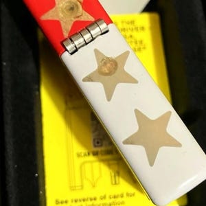 Op de afbeelding: Een Zippo aansteker met een rode bovenkant en witte behuizing, versierd met gouden sterren. De aansteker zit in een zwarte doos met een gele kaart zichtbaar, en de tekst "in USA" staat op de doos.