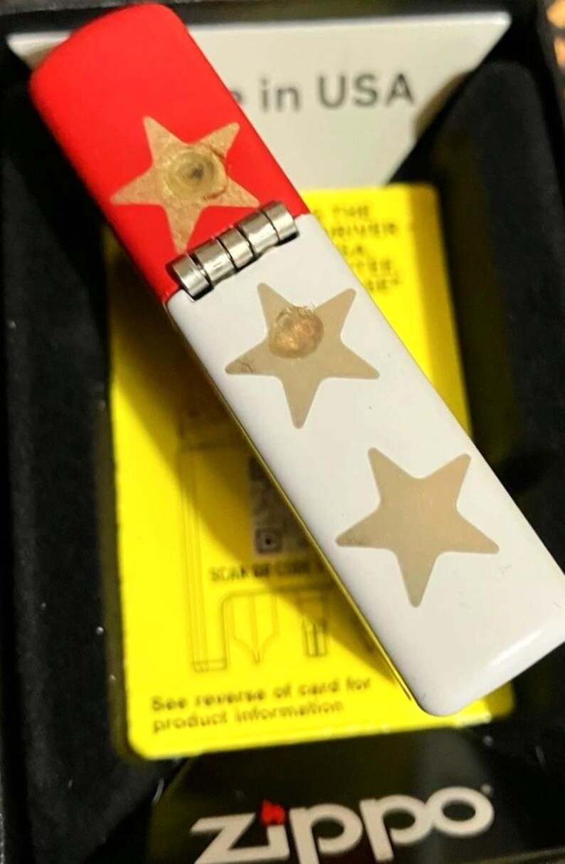 Op de afbeelding: Een Zippo aansteker met een rode bovenkant en witte behuizing, versierd met gouden sterren. De aansteker zit in een zwarte doos met een gele kaart zichtbaar, en de tekst "in USA" staat op de doos.