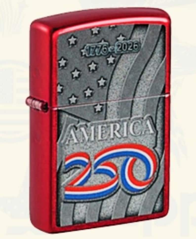 May include: A red metal lighter with a patriotic design. The lighter features a silver panel with the word "AMERICA" and the number "250" in red, white, and blue. The panel also includes stars, stripes, and the dates "1776-2026".