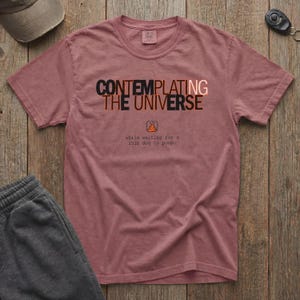 May include: A dusty rose t-shirt with the text "CONTEMPLATING THE UNIVERSE" in black and orange. Below the text is a graphic of a dog's poop and the text "while waiting for a 6.8kg dog to poop."