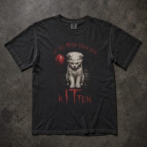 Puede incluir: Una camiseta de manga corta gris carbón con un gráfico de un gatito con maquillaje de payaso, un globo rojo y el texto WE ALL MEOW DOWN HERE KITTEN impreso en rojo. La camiseta tiene cuello redondo y un corte estándar.