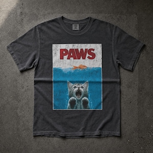 Op de afbeelding: Een antracietgrijs t-shirt met korte mouwen en een grafische print van een kat die onder water zwemt met de bek open. Boven de kat zwemt een oranje vis bij het oppervlak. De tekst PAWS staat in grote rode letters aan de bovenkant gedrukt.